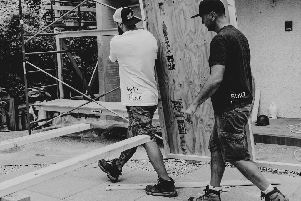 Two people carry a large sheet of plywood