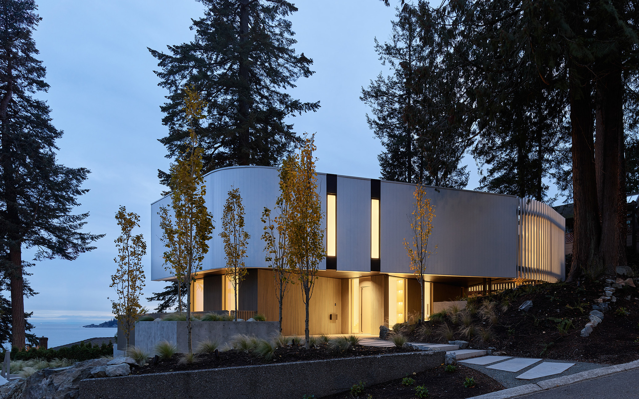 Contemporary custom home exterior at dusk with curved white cladding and forest setting overlooking the ocean