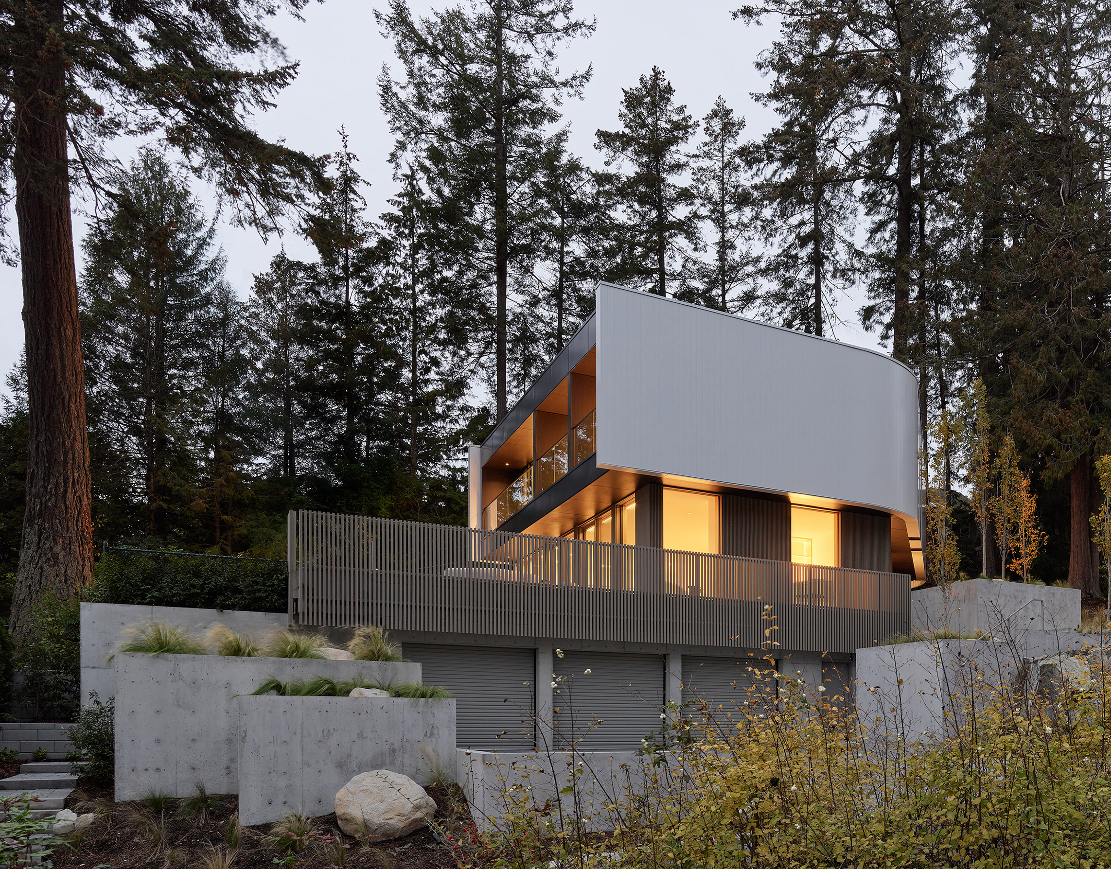 Modern cantilevered custom home with white metal cladding and concrete base surrounded by tall forest trees at dusk