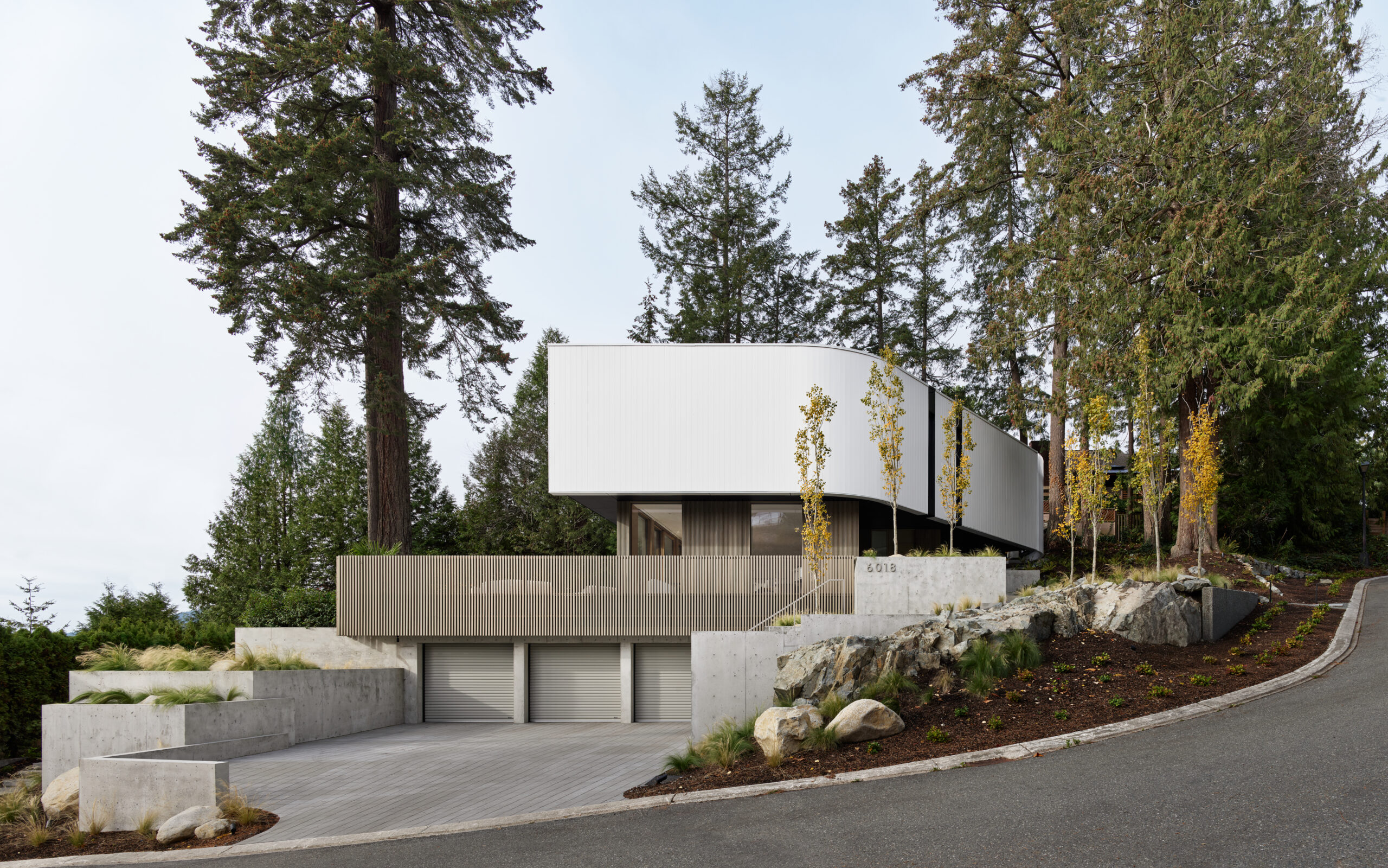 Contemporary custom hillside home with curved white cladding, concrete terraces, garages, and forest setting